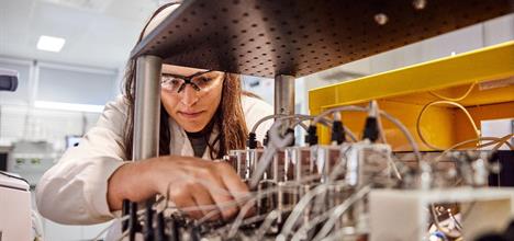 Woman in a lab wearing a lab coat and goggles while adjusting some wiring