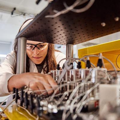 Woman in a lab wearing a lab coat and goggles while adjusting some wiring