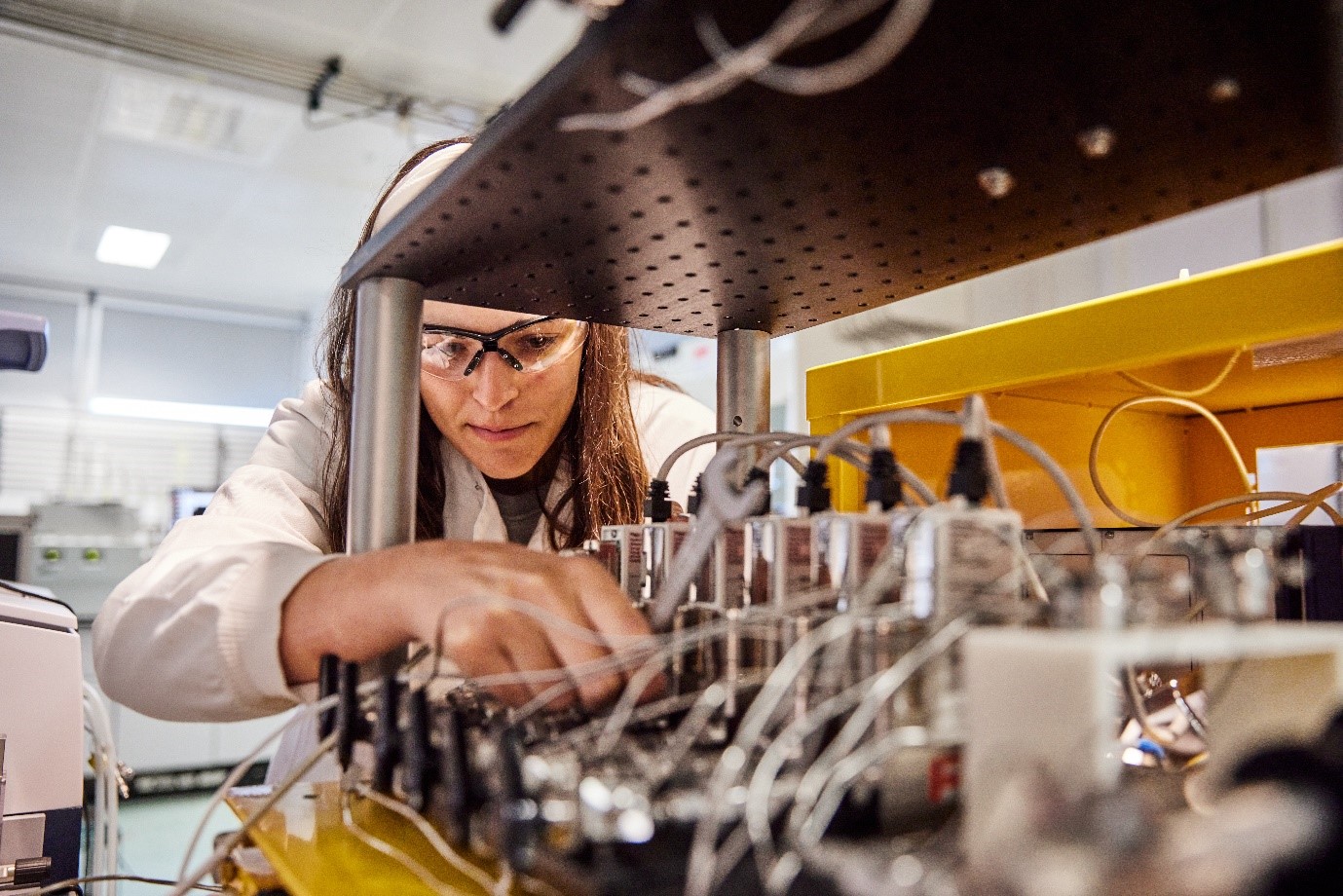 Woman in a lab wearing a lab coat and goggles while adjusting some wiring