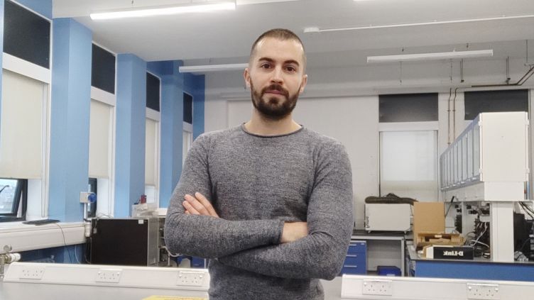 Dr Marco Amores stands confidently with arms crossed in a bright, modern laboratory. He is wearing a grey jumper and has a close-cropped haircut and beard. Behind him are lab benches, scientific equipment, and blue and white walls with large windows.