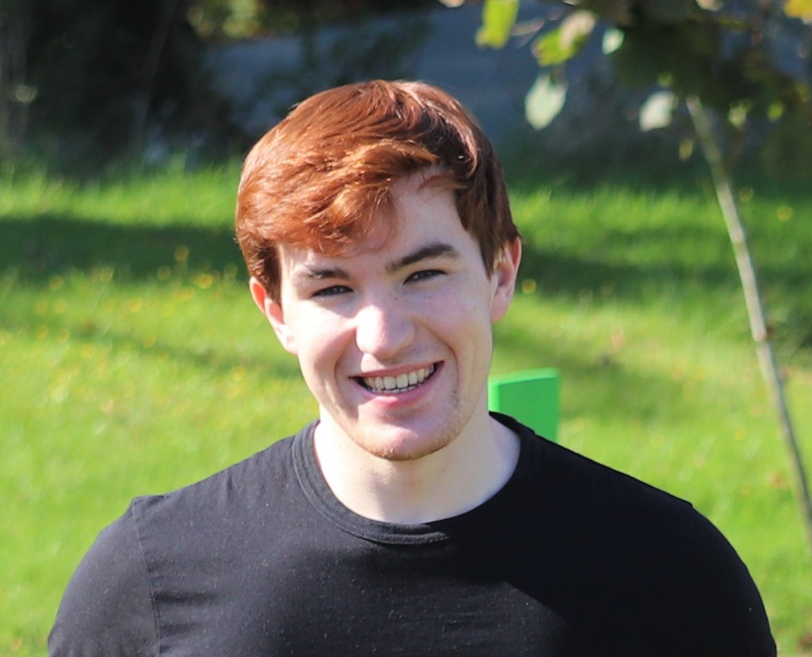 James Durant ISIS placement student at the University of Warwick. He has short red hair, and smiles outdoors on a sunny day. He is wearing a black t-shirt and is standing in front of a grassy area with a few trees and a pathway in the background.